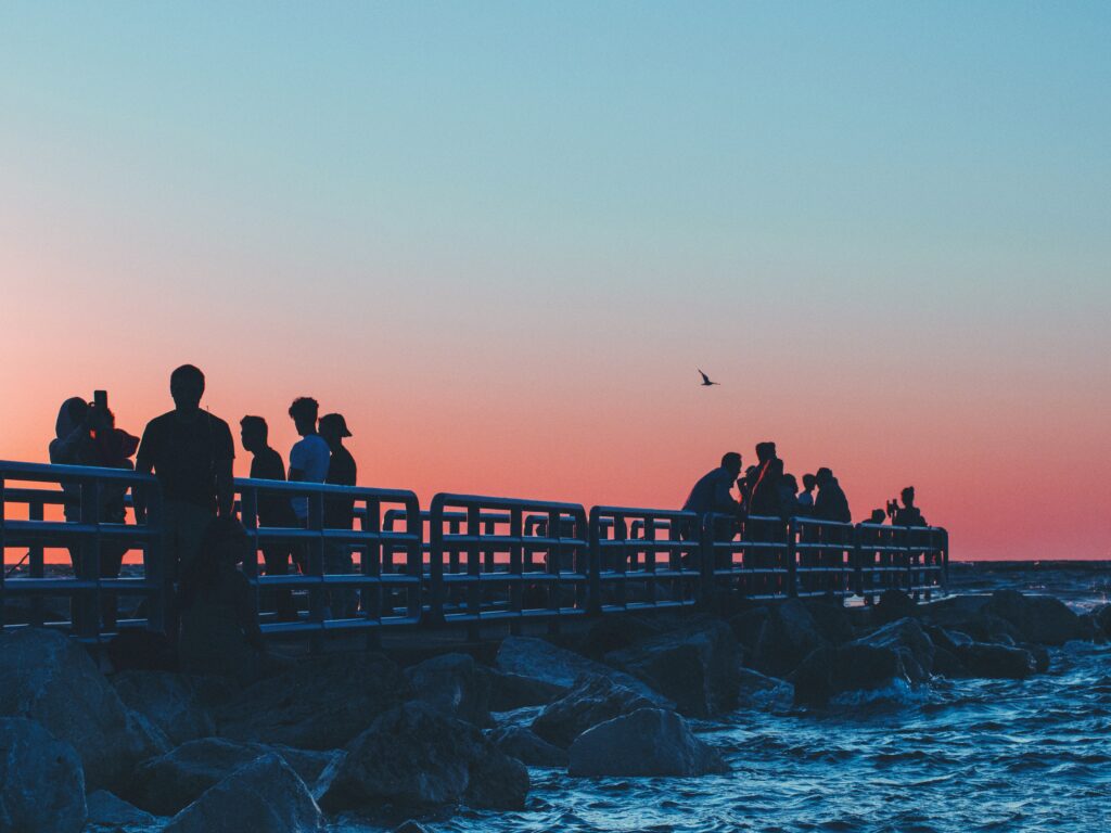 Small crowds of people stand and take pictures on a walkway over a rocky shore at twilight.