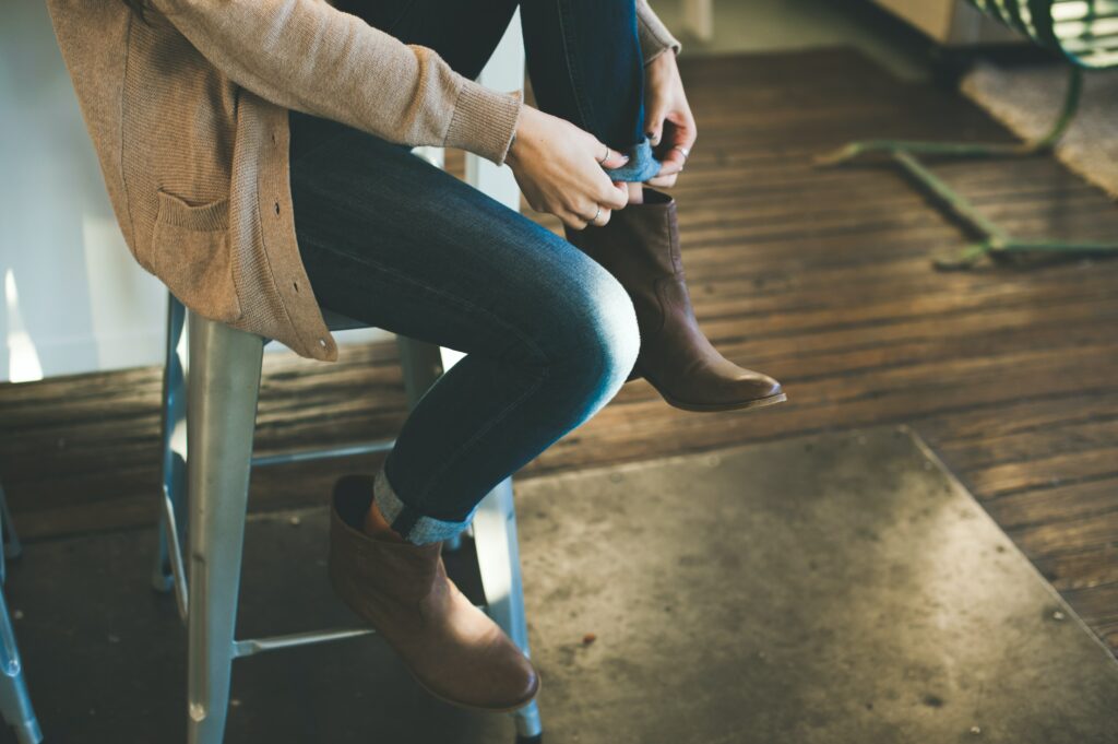 A person sits on a barstool, with only their legs in frame, as they cuff their jeans into their cowboy-style boots.